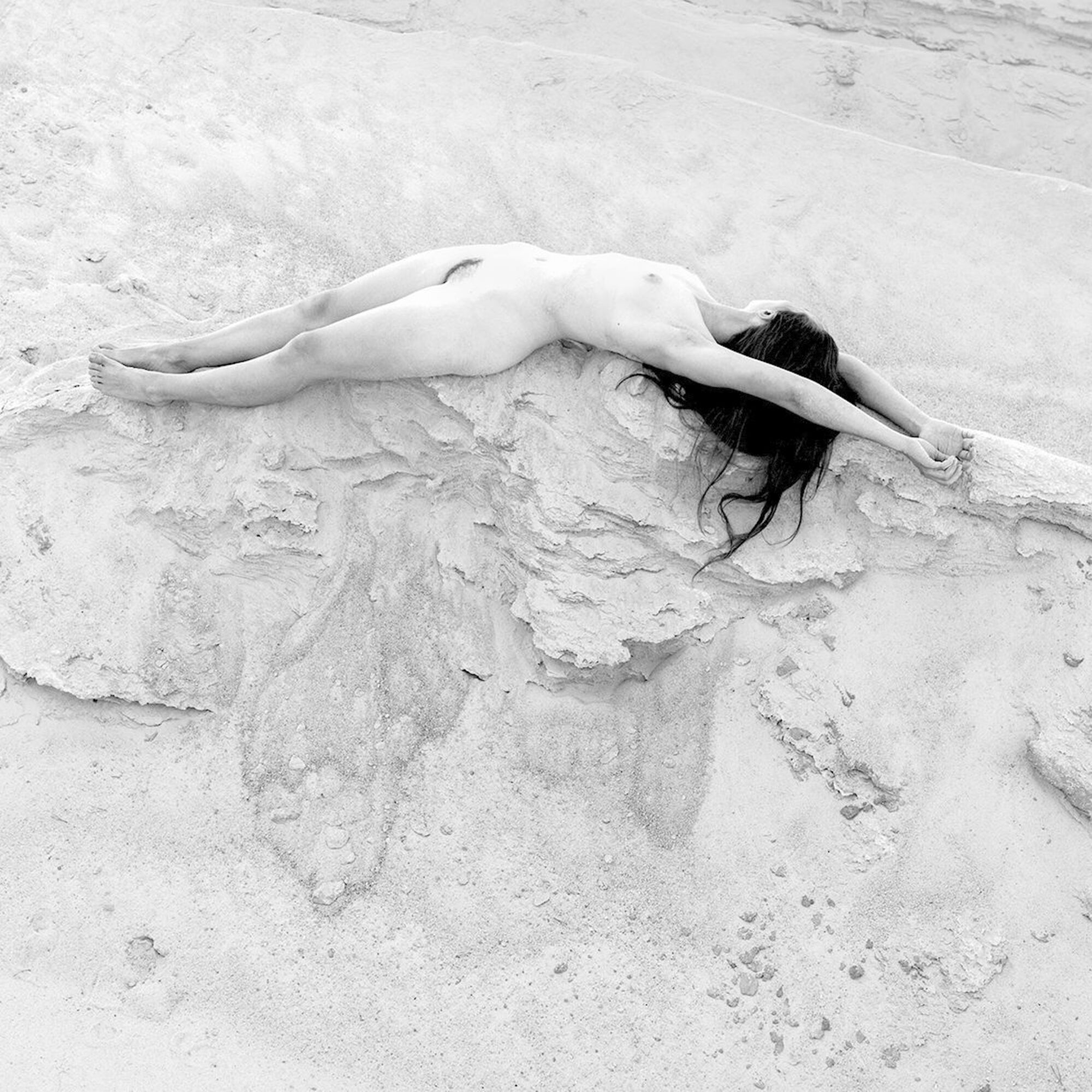 Ein schwarz-weißes Foto. Auf dem Foto ist einer Akt von einer Frau. Die Frau liegt an dem Sand.
