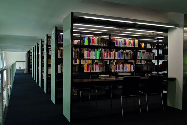 Bibliothek der Hochschule. Mehrere Bücherregale. Schwarzer Tisch mit Stühlen.
