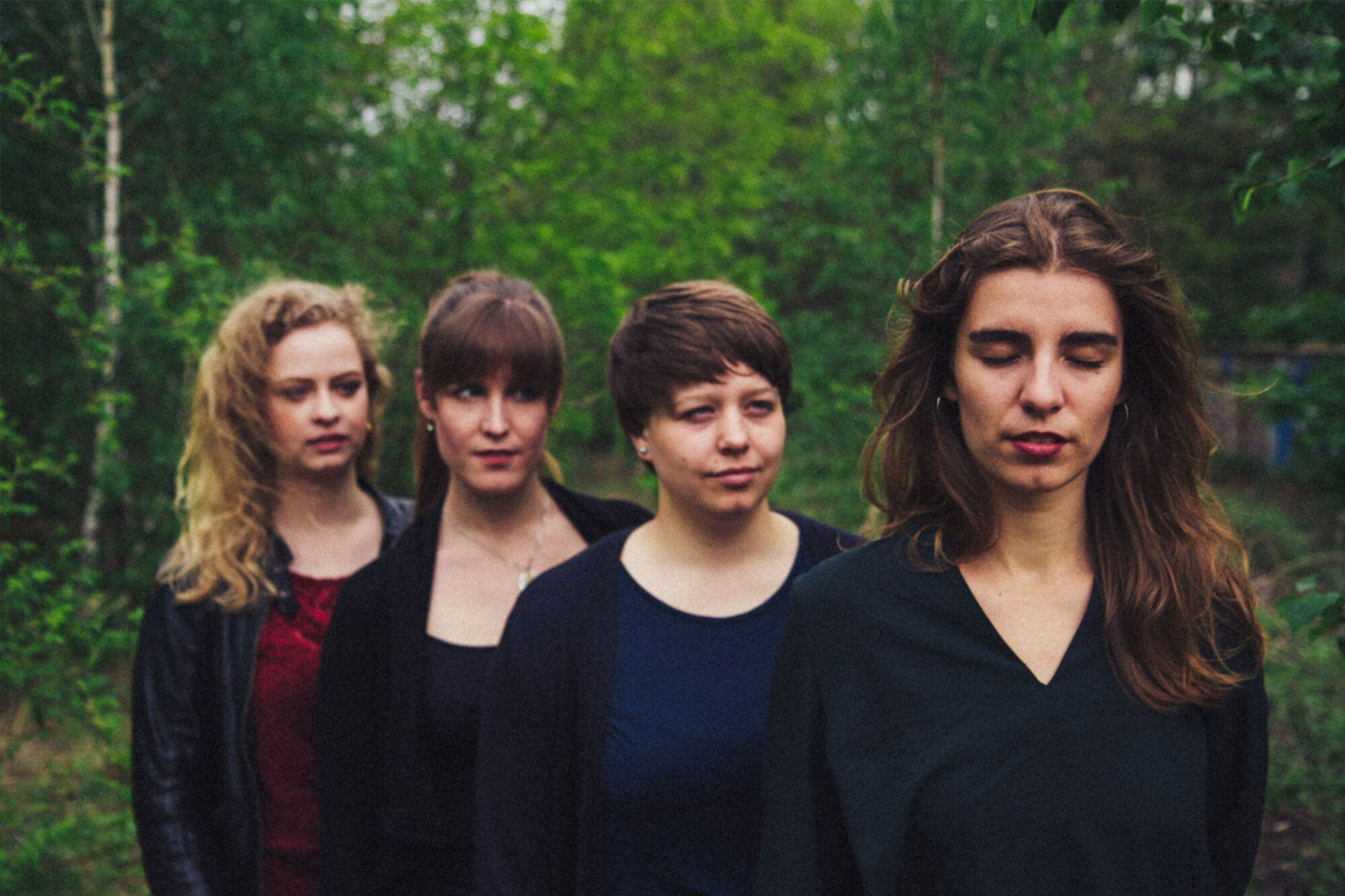 Vier Frauen stehen hintereinander im Wald. Sie sind in dunklen Farben gekleidet.