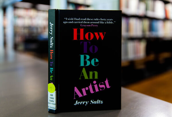 The book "How to be an artist" set on a table, in the background are library shelves.
