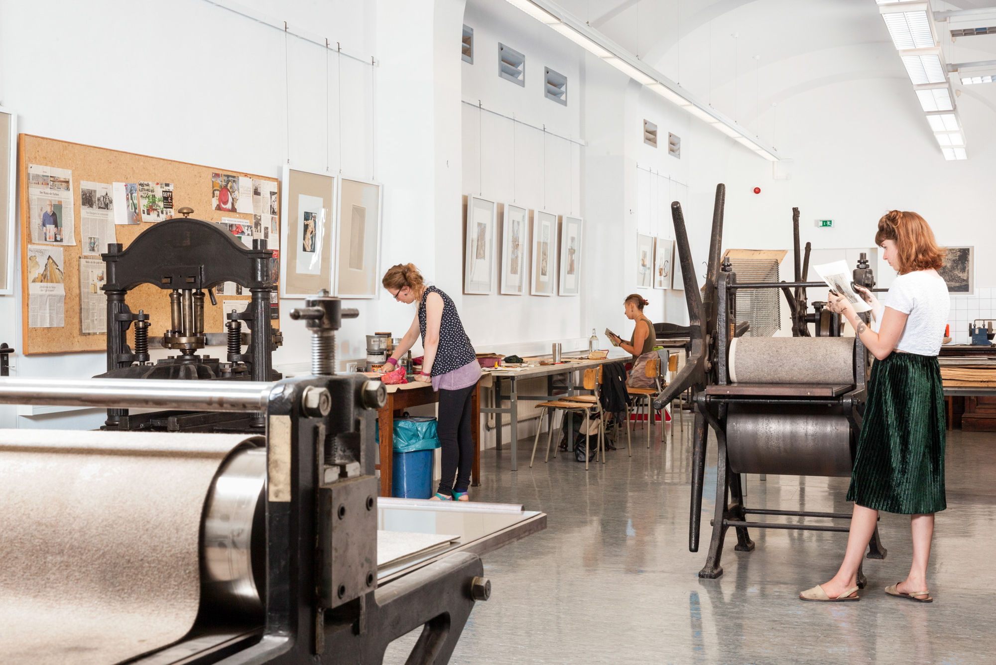 Werkstatt für Radierung. Kunstwerke auf der Wand. Arbeitstische, Druckpresse und verschiedene Werkzeuge für Radierung. Drei Studentinnen beim Arbeiten.