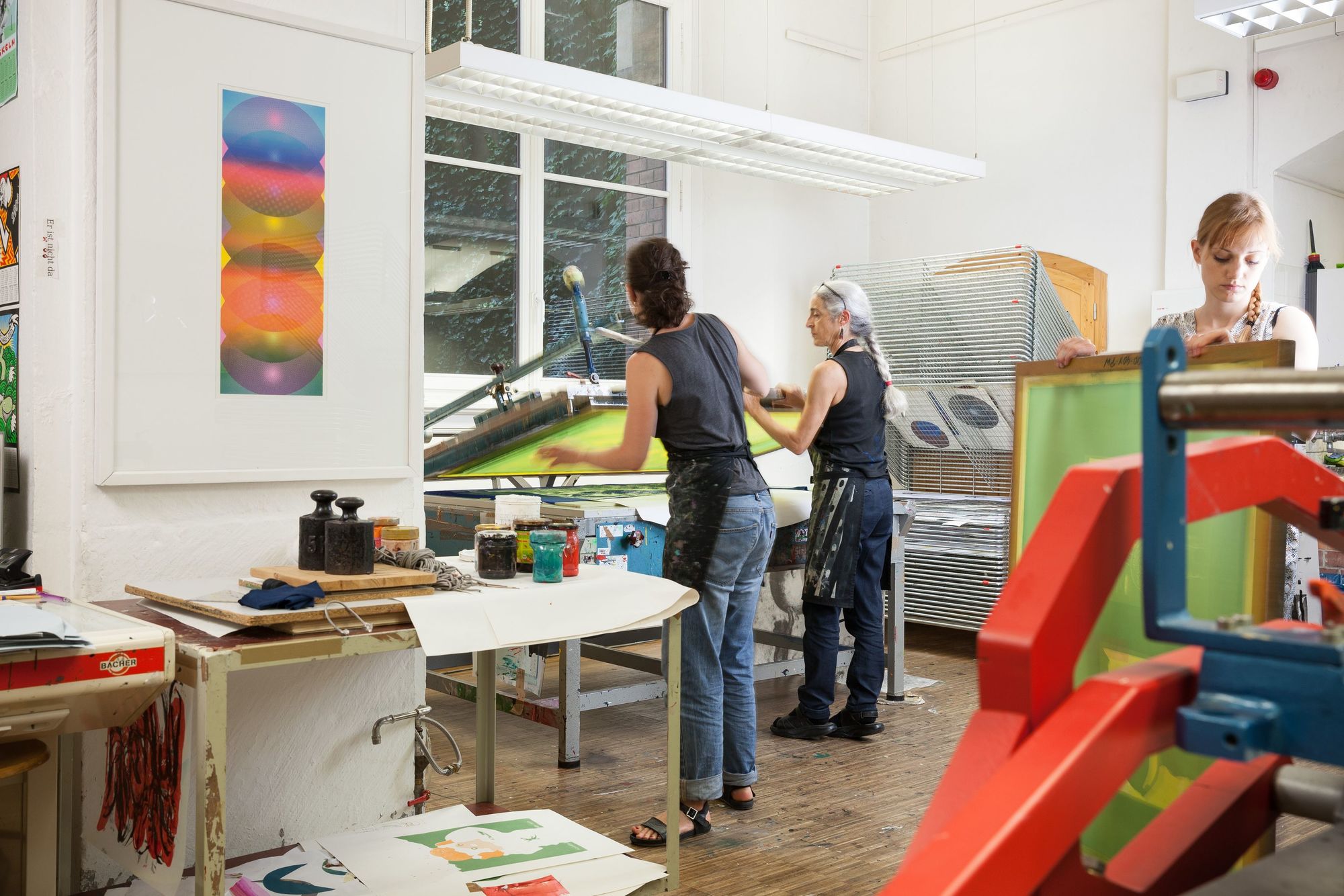 Werkstatt für Siebdruck. Siebdruckpresse, Stapeltrockner, Siebdruckplatten, verschiedene Farben und andere Werkzeuge für Siebdruck. Drei Menschen beim Arbeiten.