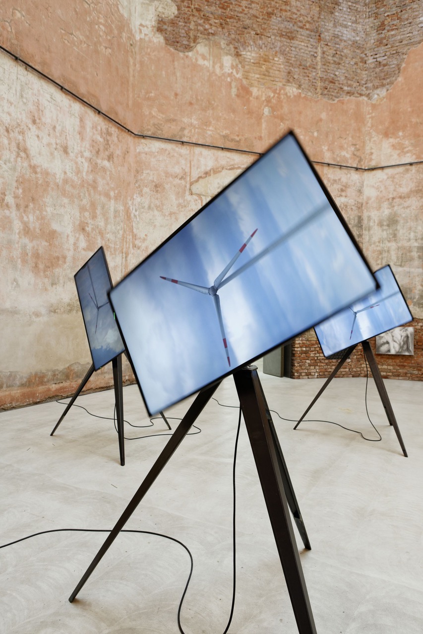 Einzelne Windrad Videoskulptur. Auf einem Dreibein ist ein großer TV angebracht, der sich dreht. Auf dem TV ist das Video eines Windrads zu sehen.