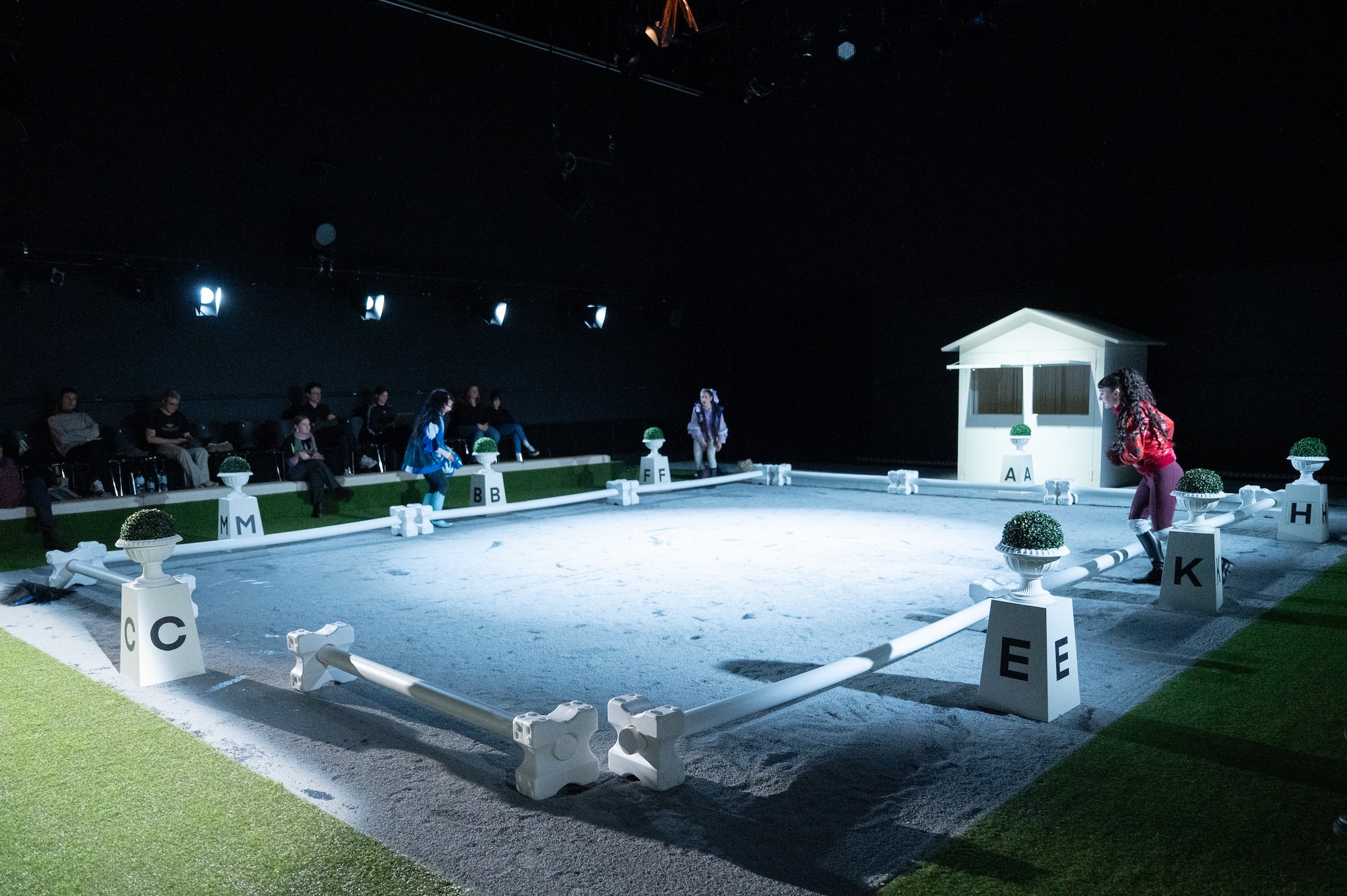 Eine ebenerdige Bühne. Sie ist gestaltet wie ein Reitplatz mit Sand und kleinen Hindernissen.