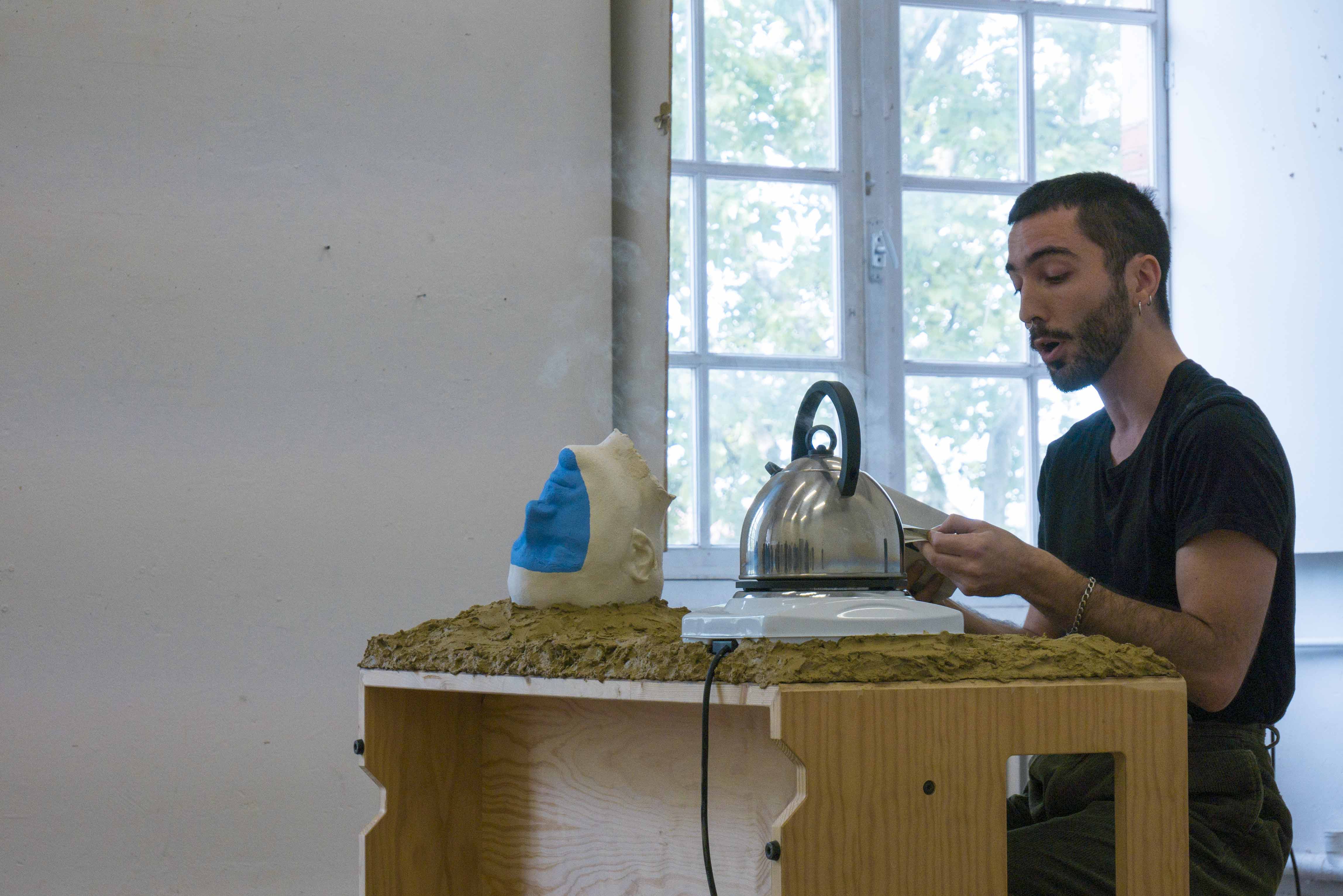 Performer verliest text an einem Tisch ausgestattet mit Kochplatte, Wasserkessel und Kunstobjekt