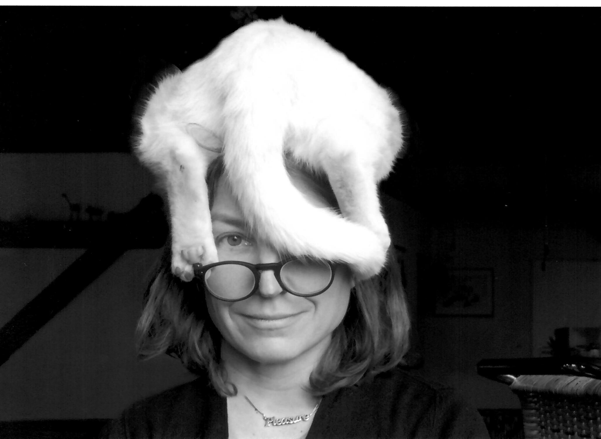 Schwarz-weiß Portrait der Künstlerin Nina Rhode, sie lächelt unter einer Hutskulptur auf ihrem Kopf hervor.