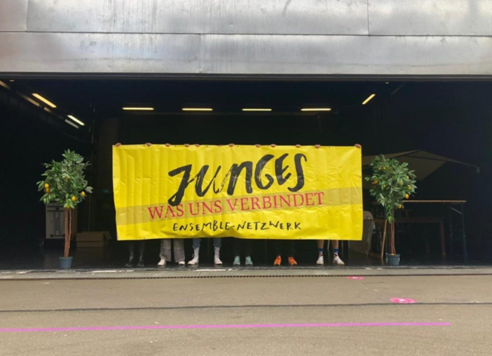 Menschen halten ein gelbes Transpi, auf dem die Worte "Junges Ensemblenetzwerk" und "Was uns verbindet" stehen.